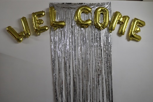 Gold foil balloons spell out the word 'WELCOME' and are arranged above a backdrop made of long, vertical metallic silver streamers against a white wall.
