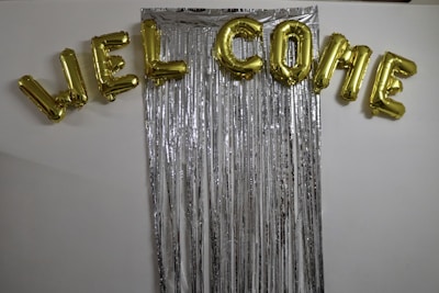 Gold foil balloons spell out the word 'WELCOME' and are arranged above a backdrop made of long, vertical metallic silver streamers against a white wall.