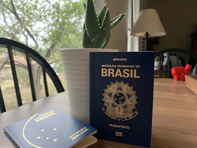 Two Brazilian passports are placed on a wooden table near a white potted plant, amidst a cozy indoor setting with a lamp and a view of greenery through the window.