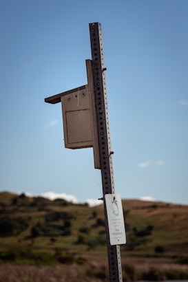 A wooden birdhouse is mounted on a metal pole against a backdrop of rolling green hills under a clear blue sky. A sign is attached to the pole, cautioning visitors not to disturb the area. The birdhouse appears weathered and part of a tranquil rural landscape.