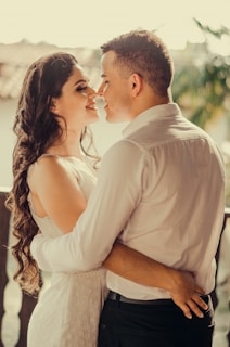 A couple stands closely together, looking into each other's eyes with smiles. The woman has long, wavy hair and is wearing a white dress, while the man is dressed in a white shirt. They are embracing each other warmly. In the background, there are blurred view of trees and rooftops.