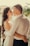 A couple stands closely together, looking into each other's eyes with smiles. The woman has long, wavy hair and is wearing a white dress, while the man is dressed in a white shirt. They are embracing each other warmly. In the background, there are blurred view of trees and rooftops.
