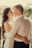A couple stands closely together, looking into each other's eyes with smiles. The woman has long, wavy hair and is wearing a white dress, while the man is dressed in a white shirt. They are embracing each other warmly. In the background, there are blurred view of trees and rooftops.