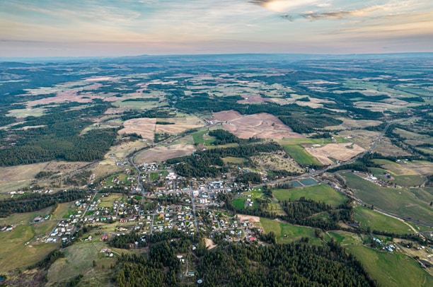 Aerial view of a rural landscape featuring a small town with buildings scattered among green fields, patches of forests, and agricultural lands. The terrain stretches into the distance with a few hills visible on the horizon under a partially cloudy sky.