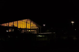 A modern building with a slanted roof is illuminated from the inside, casting a warm orange glow through large glass windows. The surrounding area is dark, with two streetlights providing minimal light to the exterior.