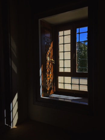 Light filters through a decorative wooden window with metal latticework, casting intricate shadows on the dark interior walls and floor. Outside, a clear blue sky is visible.
