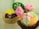 Two cupcakes are decorated with intricate fondant designs. One cupcake features a bright yellow fondant flower with a silver bead at its center, sitting on a chocolate base in a gold paper liner. The other cupcake, displayed in a brown paper liner, is adorned with pink fondant roses and a pastel green fondant leaf.