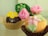 Two cupcakes are decorated with intricate fondant designs. One cupcake features a bright yellow fondant flower with a silver bead at its center, sitting on a chocolate base in a gold paper liner. The other cupcake, displayed in a brown paper liner, is adorned with pink fondant roses and a pastel green fondant leaf.