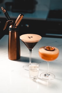 On a white marble countertop, there is a tall copper-colored cream whipper next to a martini glass filled with a creamy espresso cocktail. Beside it is another cocktail in a coupe glass, topped with a halved passion fruit. In the foreground, there is a small shot glass filled with a clear liquid.