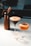 On a white marble countertop, there is a tall copper-colored cream whipper next to a martini glass filled with a creamy espresso cocktail. Beside it is another cocktail in a coupe glass, topped with a halved passion fruit. In the foreground, there is a small shot glass filled with a clear liquid.