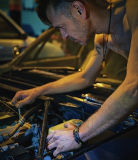 A person is working on a car engine, focusing intently as they use both hands to adjust or repair parts under the open hood. The scene conveys a sense of concentration and dedication, with ambient lighting highlighting the muscles and details of the engine components.