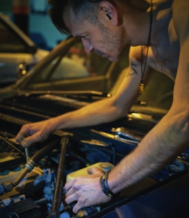 A person is working on a car engine, focusing intently as they use both hands to adjust or repair parts under the open hood. The scene conveys a sense of concentration and dedication, with ambient lighting highlighting the muscles and details of the engine components.
