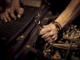 Hands working on a car engine, one grasping a wrench while the other steadies radiator fins. The scene is focused on the forearms and tools, highlighting mechanical components.