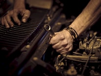Hands working on a car engine, one grasping a wrench while the other steadies radiator fins. The scene is focused on the forearms and tools, highlighting mechanical components.