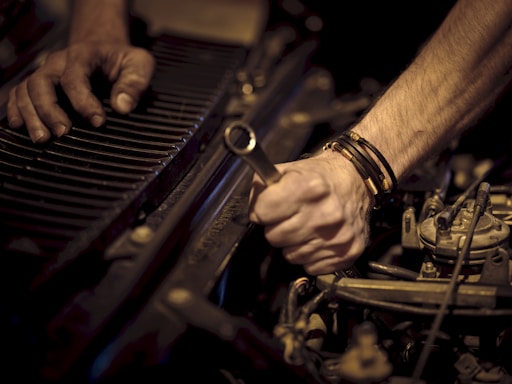 Hands working on a car engine, one grasping a wrench while the other steadies radiator fins. The scene is focused on the forearms and tools, highlighting mechanical components.