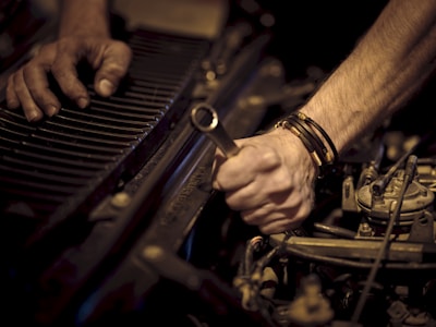 Hands working on a car engine, one grasping a wrench while the other steadies radiator fins. The scene is focused on the forearms and tools, highlighting mechanical components.