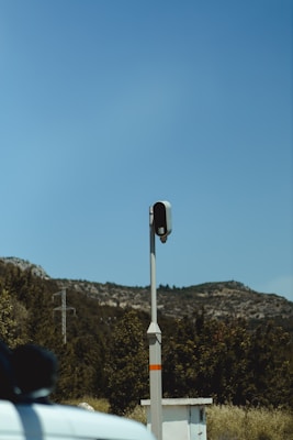 A traffic camera mounted on a tall pole with a mountainous landscape in the background. The sky is clear and blue, and there are trees and vegetation in the lower part of the image. Part of a white vehicle is visible in the foreground.