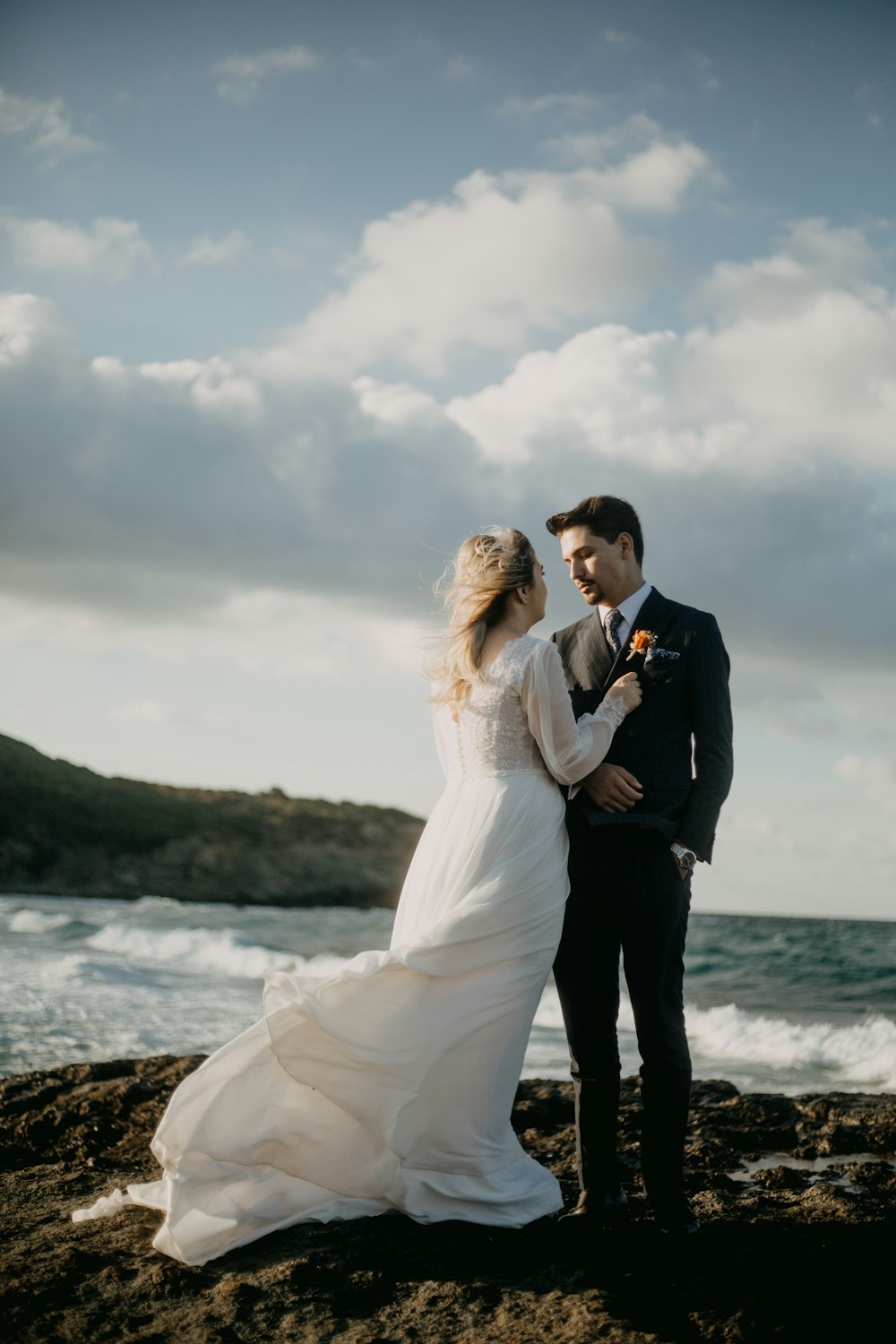 homme en costume noir embrassant femme en robe de mariée blanche sur le bord de mer pendant la journée