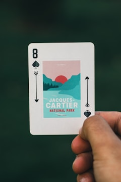 A playing card is held between two fingers against a blurred green background. It features a design for Jacques-Cartier National Park with an artistic illustration of mountains and a sun. The card is marked as the eight of spades.