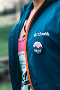 A close-up of a person wearing a blue Columbia jacket with an orange zipper. A button with the text 'Kootenay National Park' is pinned to the jacket. Underneath the jacket, a pink garment featuring some text is visible.