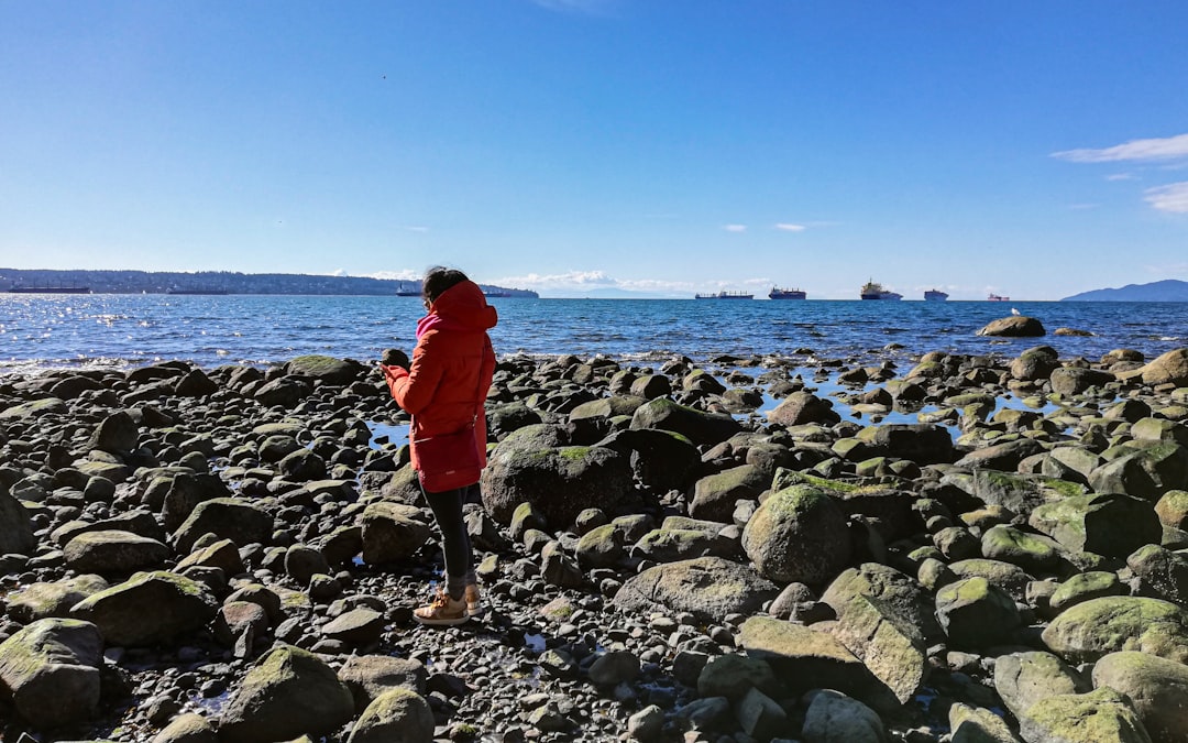 woman in red jacket and black pants standing on rocky shore during daytime