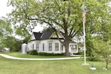 A charming, white, vintage house with black roof shingles sits nestled among lush green trees. A well-maintained lawn surrounds the house, and an American flag flies on a flagpole. The house features a prominent front porch and a pathway leading to the entrance.