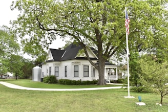 A charming, white, vintage house with black roof shingles sits nestled among lush green trees. A well-maintained lawn surrounds the house, and an American flag flies on a flagpole. The house features a prominent front porch and a pathway leading to the entrance.