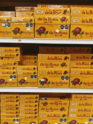 Shelves stacked with multiple boxes of 'de la Rosa' products, featuring a prominent rose logo and clear branding on yellow packaging.