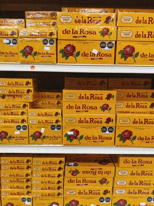 Shelves stacked with multiple boxes of 'de la Rosa' products, featuring a prominent rose logo and clear branding on yellow packaging.