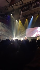 A live concert scene with a crowd watching a band perform on stage. Bright stage lights in various colors illuminate the performers, and a large screen in the background adds visual effects. The atmosphere is lively and energetic.