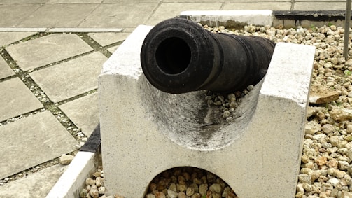A historic cannon is mounted on a simple, white concrete base surrounded by small stones. The setting appears to be outdoor, with a tiled pavement visible in the background. The cannon has a dark, weathered surface, indicating its age and historical significance.