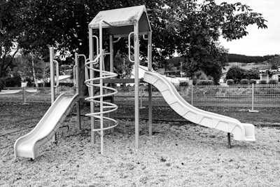 A playground structure featuring two slides and a spiral ladder, set on a gravel surface. The playground is situated in an outdoor area with trees in the background, and a metal fence encloses the area.