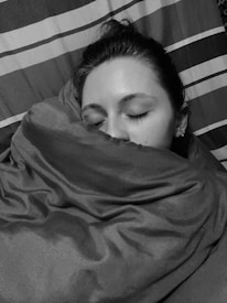 A person with closed eyes rests their head on a pillow while wrapped in a blanket. The bedding features striped patterns, and the image appears in grayscale.