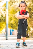 A young child with dark hair tied up stands confidently outdoors with arms crossed. They are wearing a black outfit with a red scarf featuring a black star. Bright sunshine filters through trees in the background, creating a warm, illuminated atmosphere.