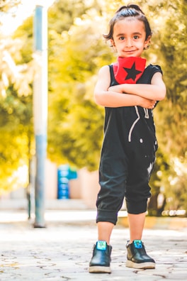 A young child with dark hair tied up stands confidently outdoors with arms crossed. They are wearing a black outfit with a red scarf featuring a black star. Bright sunshine filters through trees in the background, creating a warm, illuminated atmosphere.