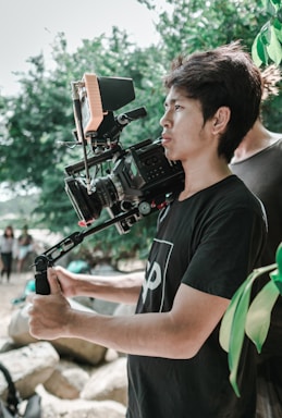 A person is holding a professional video camera mounted on a stabilizer. They are outdoors, surrounded by greenery and rocks. The individual appears focused on capturing a scene.