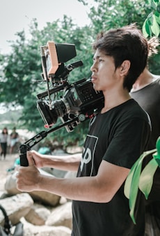 A person is holding a professional video camera mounted on a stabilizer. They are outdoors, surrounded by greenery and rocks. The individual appears focused on capturing a scene.