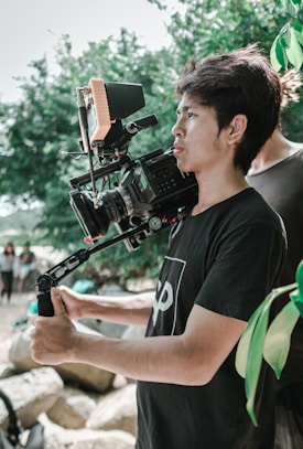 A person is holding a professional video camera mounted on a stabilizer. They are outdoors, surrounded by greenery and rocks. The individual appears focused on capturing a scene.