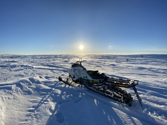 A snowmobile is parked on a vast, snow-covered landscape under a clear blue sky with the sun low on the horizon. The sunlight casts long shadows on the snow, creating a crisp and serene winter ambiance.