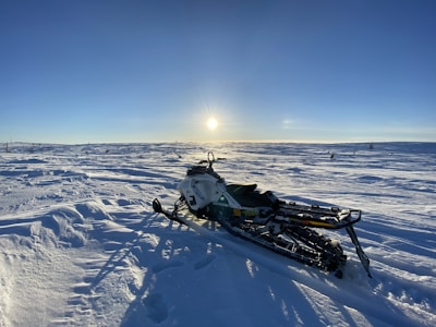 A snowmobile is parked on a vast, snow-covered landscape under a clear blue sky with the sun low on the horizon. The sunlight casts long shadows on the snow, creating a crisp and serene winter ambiance.