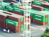 Surveyor inspecting cargo containers at a busy port under green and white company banners.
