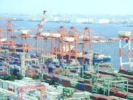 A busy shipping port with numerous cargo containers arranged in stacks and several large cranes loading and unloading ships. The cranes are orange and white, and there is a body of water in the background with ships and a city skyline in the distance.