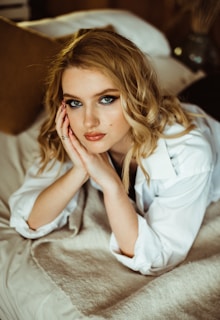 A woman with blonde wavy hair and striking blue eyes, wearing a white shirt, is lying on a bed covered with a light-colored blanket. She rests her chin on her hands and looks directly at the camera with a calm expression. The setting appears cozy and intimate, with warm neutral tones in the background.