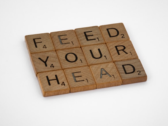 Wooden Scrabble tiles are arranged in a neat grid spelling out the phrase 'FEED YOUR HEAD.' Each tile displays a letter alongside a small number indicating its point value in the game.