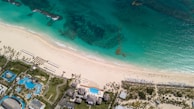 Aerial view of a tropical beach with clear turquoise water meeting a pristine sandy shore. Sun loungers and umbrellas are neatly arranged along the beach. Inland, a resort area features several pools with surrounding palm trees and landscaped gardens.