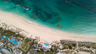 Aerial view of a tropical beach with clear turquoise water meeting a pristine sandy shore. Sun loungers and umbrellas are neatly arranged along the beach. Inland, a resort area features several pools with surrounding palm trees and landscaped gardens.