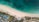 Aerial view of a tropical beach with clear turquoise water meeting a pristine sandy shore. Sun loungers and umbrellas are neatly arranged along the beach. Inland, a resort area features several pools with surrounding palm trees and landscaped gardens.