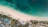 Aerial view of a tropical beach with clear turquoise water meeting a pristine sandy shore. Sun loungers and umbrellas are neatly arranged along the beach. Inland, a resort area features several pools with surrounding palm trees and landscaped gardens.
