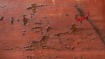 A textured brown surface features peeling paint and rust, with patches of darker corrosion scattered across the area. A small strip of red tape is placed in the upper right, contrasting with the rustic tones.