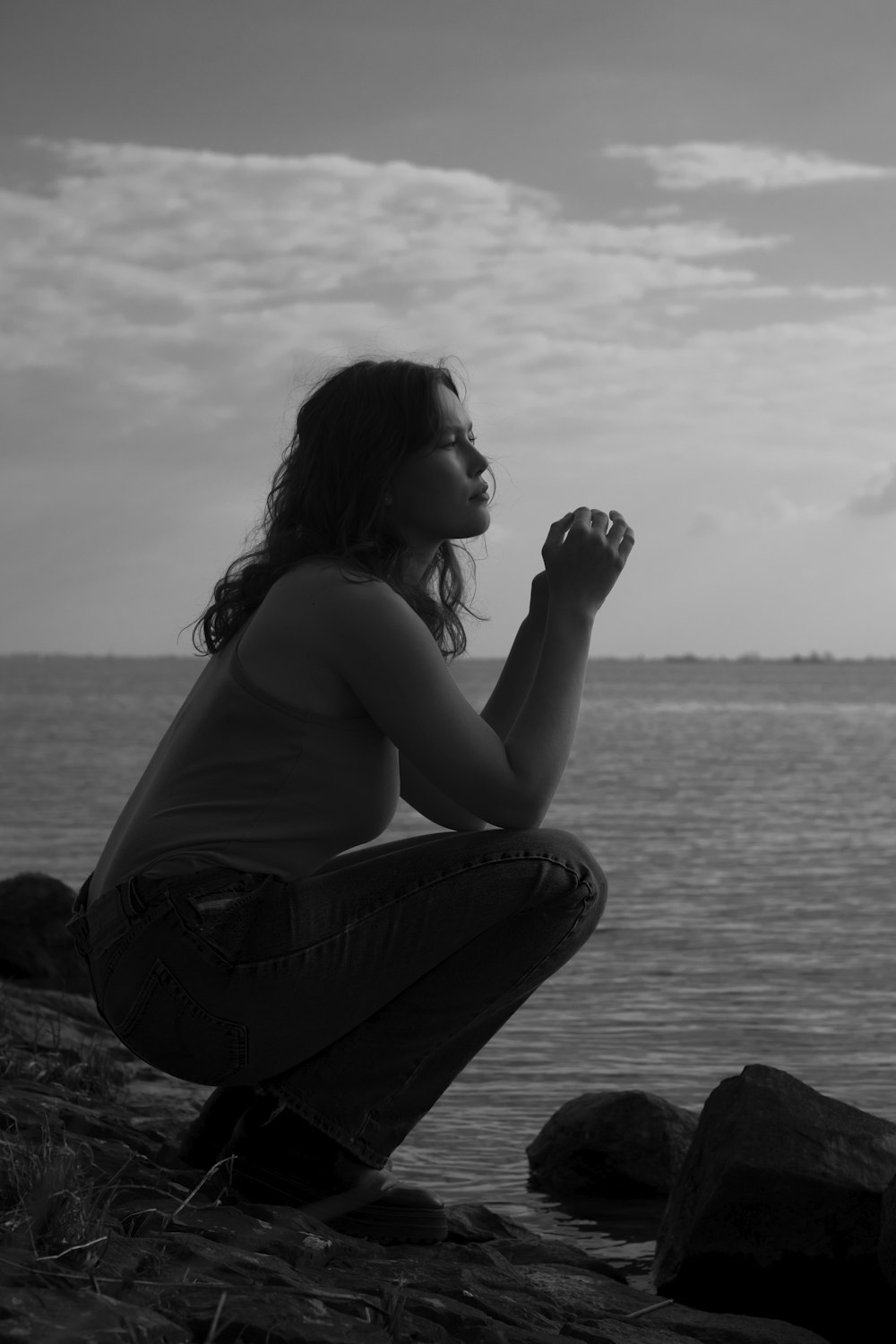 woman in black tank top and blue denim jeans sitting on rock near body of water